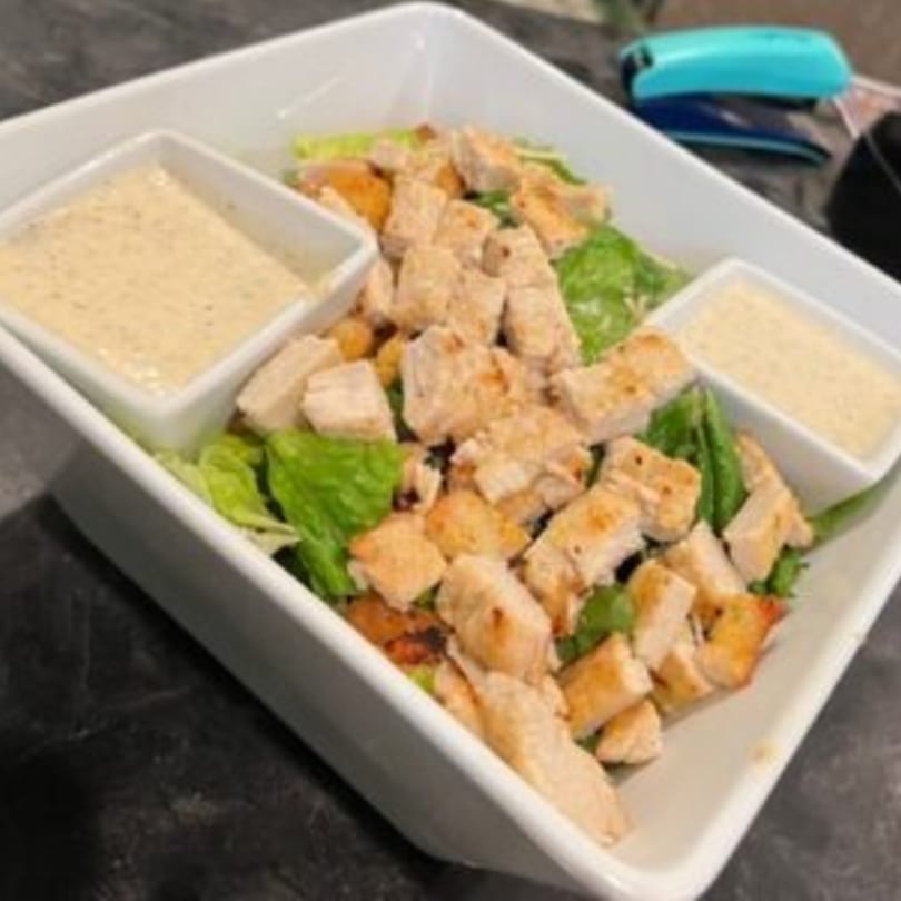 Chicken Caesar salad with two small bowls of dressing in a white rectangular dish.
