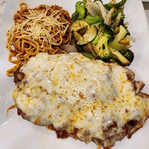 Chicken parmesan with melted cheese, a side of spaghetti, and roasted vegetables on a white plate.