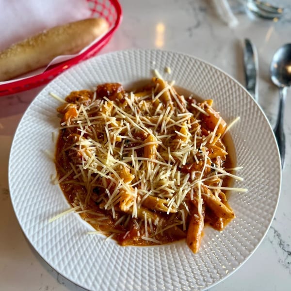 A plate of penne pasta with red sauce and shredded parmesan cheese, with a breadstick in a red basket.