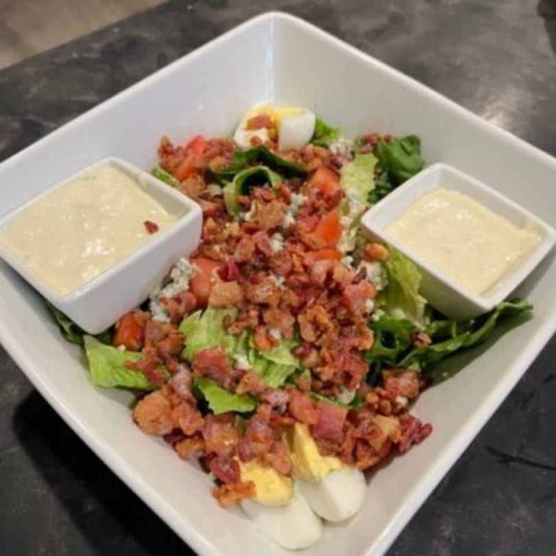 A square white bowl filled with a salad of greens, bacon, tomatoes, eggs, and two side dressings.