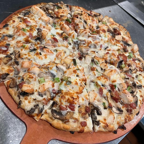 A freshly baked La Bella Vita pizza with chicken, mushrooms, bacon, and green onions on a pizza peel.
